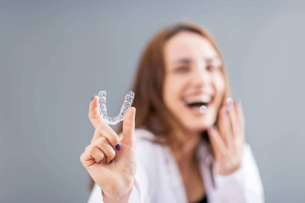 A woman holding up a clear aligner and smiling.