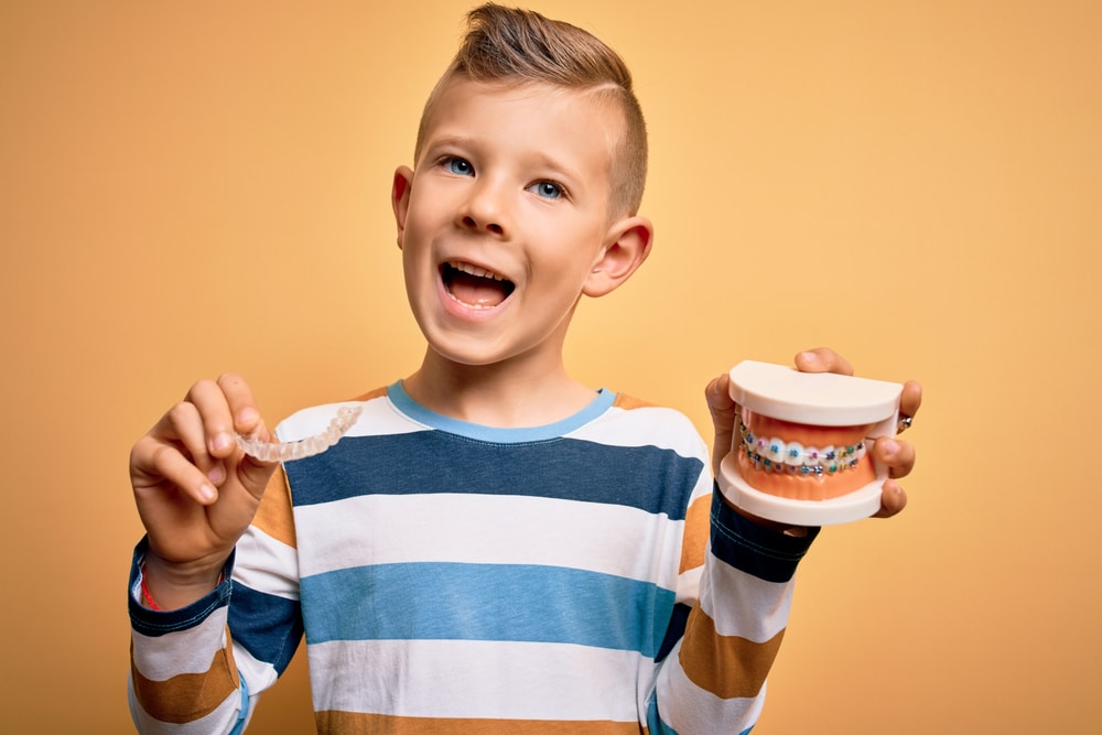 Boy in striped shirt holding a clear aligners and a model of teeth with multicolor braces.