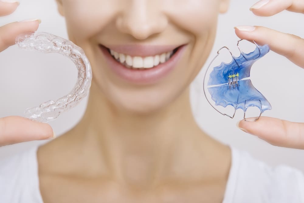 Close up of a woman smiling and holding a clear retainer and a Hawley retainer.