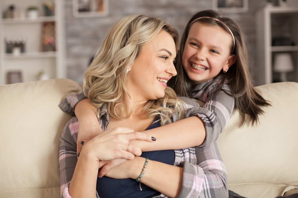 Mom and daughter with braces embracing on a cream couch.