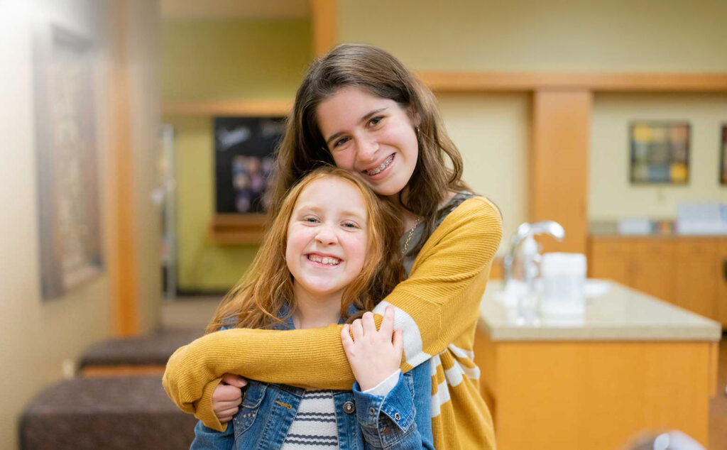 In a cheerful dental office, a teenager hugs a little girl, conveying love and reassurance during a dental visit.