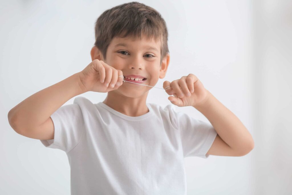 Child in a white t-shirt smiling and holding dental floss wrapped around their index fingers