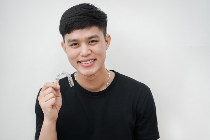 Close Up Young Asian Man Smiling With Hand Holding Dental Aligner Retainer (invisible) At White Wall Background Of Dental Clinic For Beautiful Teeth Treatment
